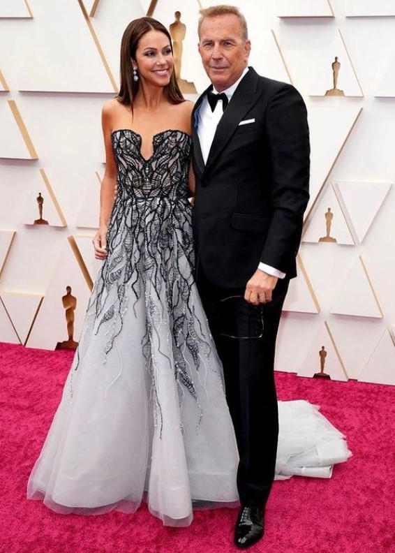 Kevin Costner wearing a black suit and Christine Baumgartner wearing a grey gown for an event.