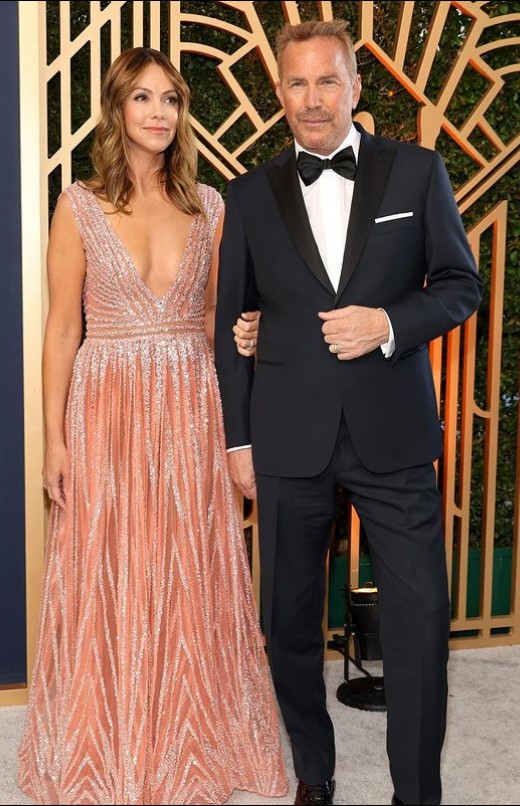 Kevin Costner wearing a black suit and white shirt and Christine Baumgartner wearing a nude colored gown.