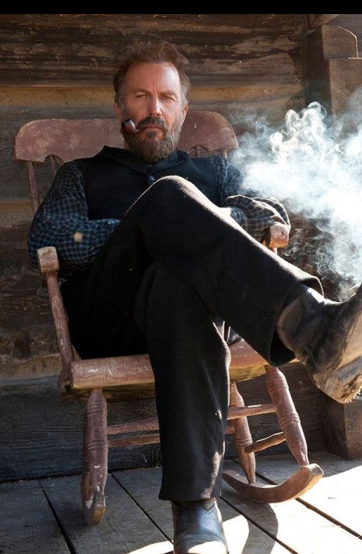 Kevin Costner wearing a black suit sitting on a wooden chair.