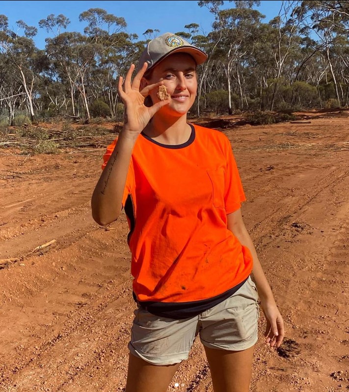 Tyler Mahoney Holding piece of gold. She is wearing an orange t shirt and brown shorts and cap.