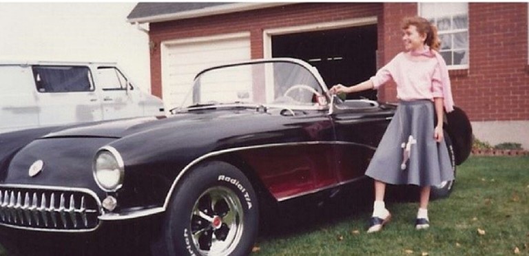 Charity Kindig posing with 1957 Corvette wearing a grey skirt and a light pink top.
