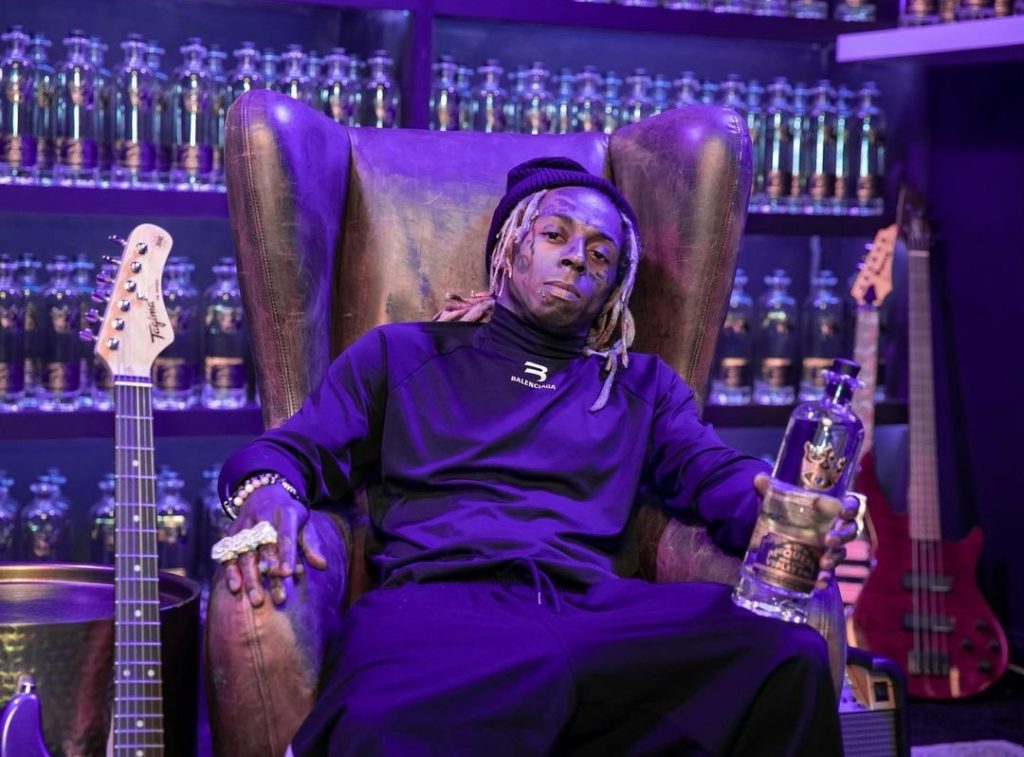Lil Wayne wearing all black clothes sitting on a chair holding a bottle in his hand.
