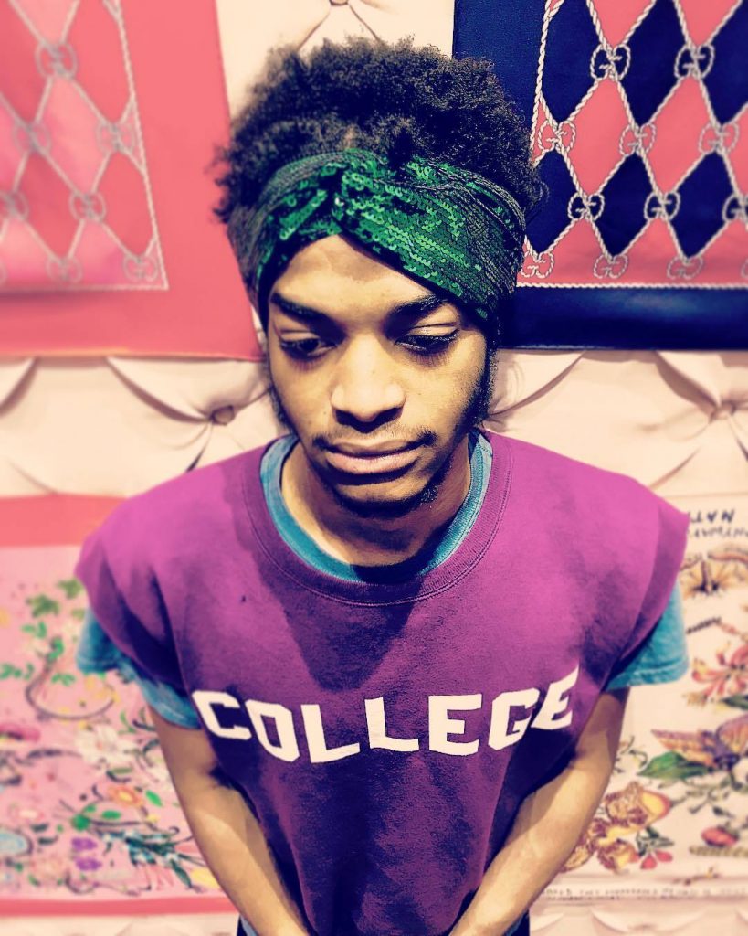 Seven wearing a green hair bandana and purple colored tee