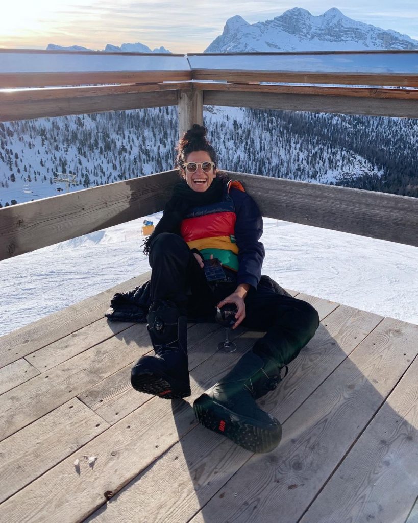 Elena wearing warm clothes in black color sitting on a wooden floor and holding a glass of red wine. She is in the middle of a scenic view covered with snow.