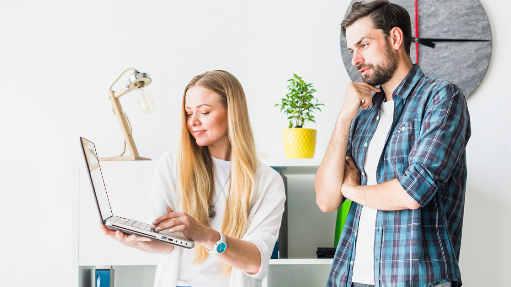 A girl wearing a white dress is showing something on her laptop to a boy wearing white tee and blue checked shirt.