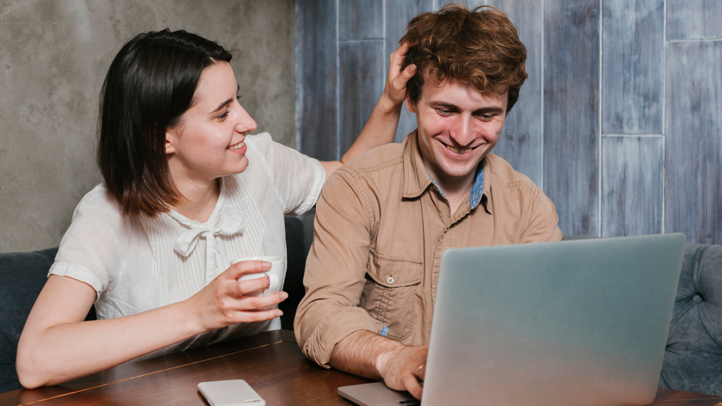 A girl wearing white top and a boy wearing brown shirt are laughing and talking. The girl is touching his hair.