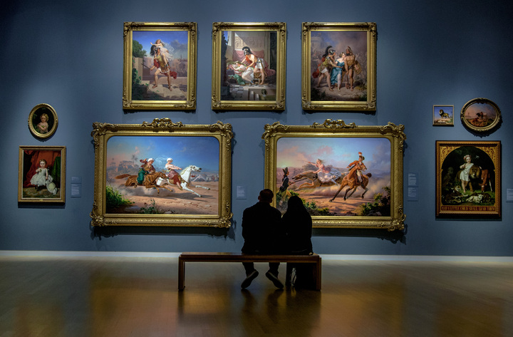 A couple sitting on the bench watching paintings in museum.
