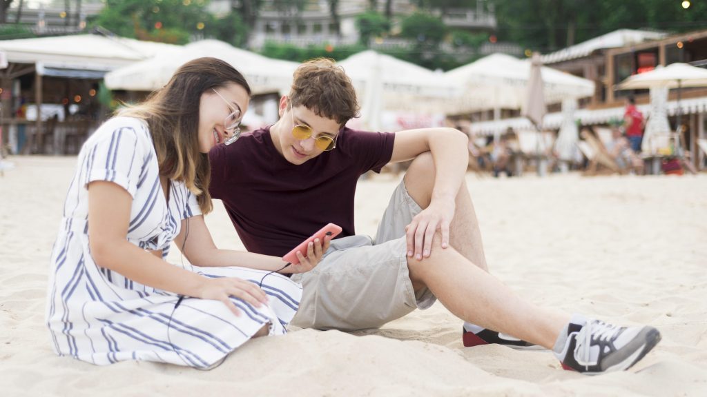 teenagers-relaxing-together at beach watching on phone.