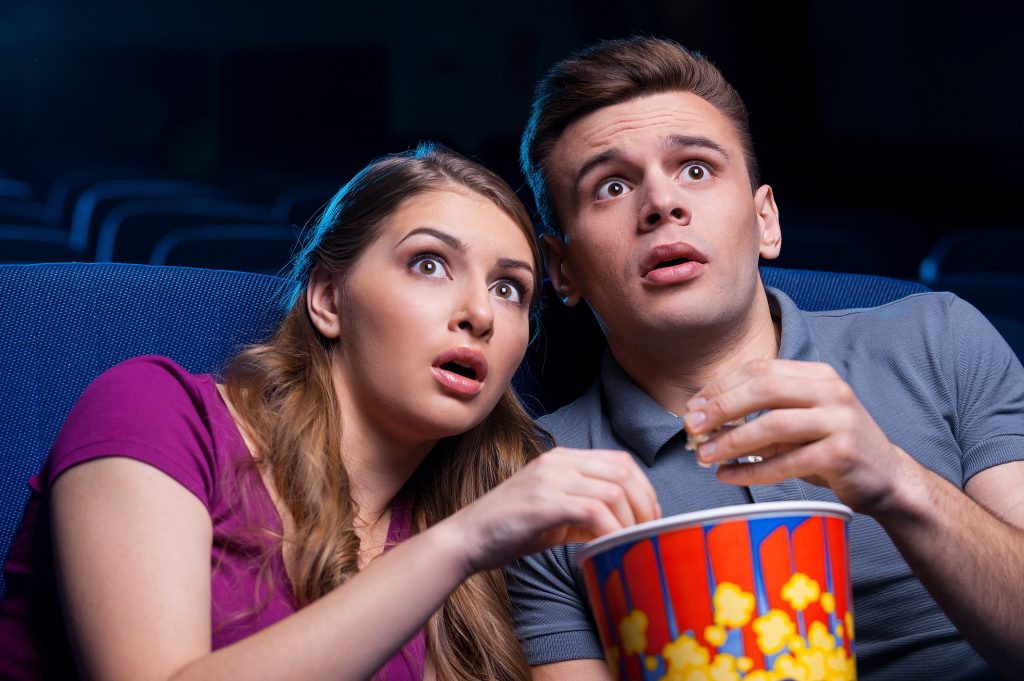 Shocked young couple eating popcorn and watching movie together while sitting at the cinema.